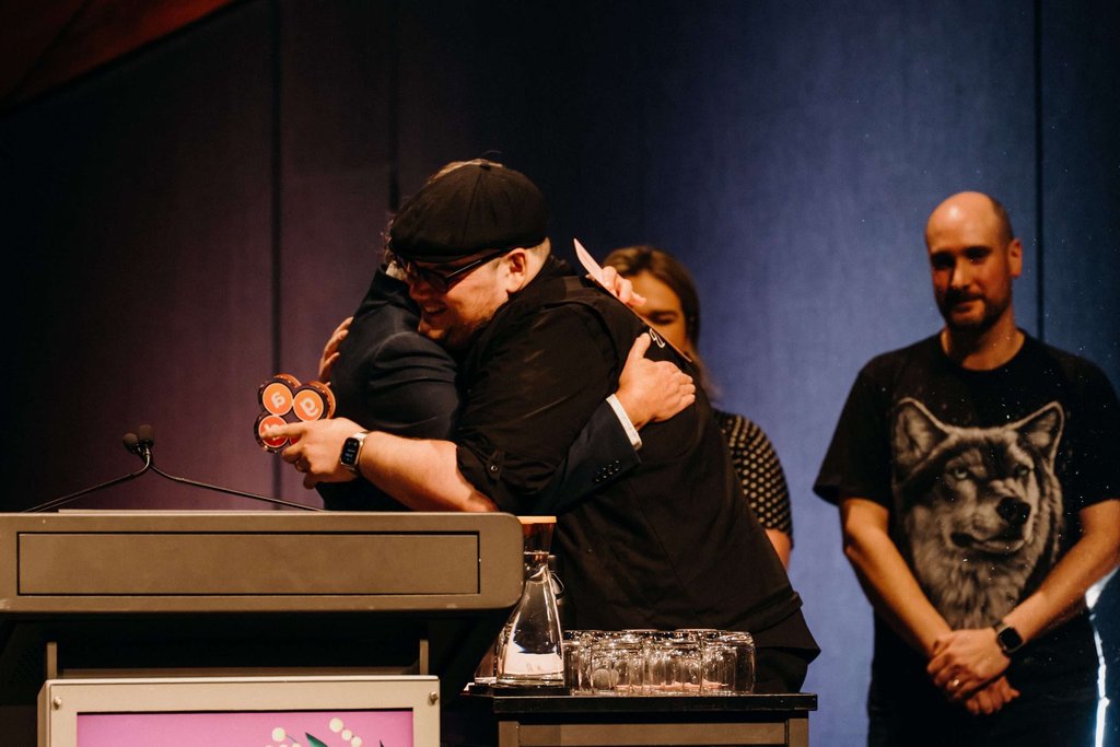 Paris hugging Tony Reed on stage beside the lectern, holding the award, with two people standing behind them.